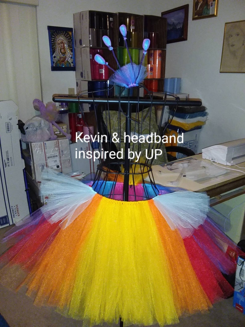 May include: A multi-colored tulle tutu skirt with a rainbow pattern. The skirt is on a mannequin and has a white, yellow, orange, red, and purple layers. The text "Kevin & headband inspired by UP" is visible in the image.