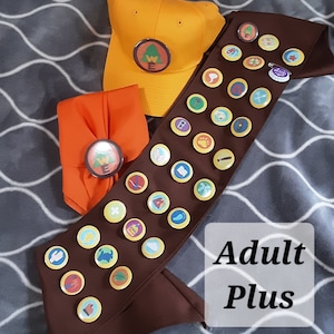 Peut inclure: Une ceinture marron avec une variété de badges colorés, une casquette jaune avec un logo d'arbre vert et une ceinture orange assortie avec un logo similaire. Le texte "Adult Plus" est visible sur l'image.