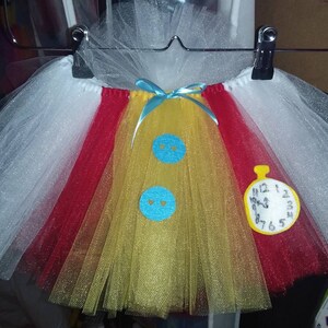 May include: A white, red, and yellow tulle tutu with blue felt buttons and a yellow felt pocket watch with black numbers. The pocket watch says "12 1 2 3 4 5 6 7 8 9 10 11 12".