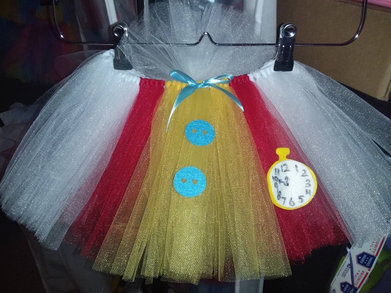 May include: A white, red, and yellow tulle tutu with blue felt buttons and a yellow felt pocket watch with black numbers. The pocket watch says "12 1 2 3 4 5 6 7 8 9 10 11 12".