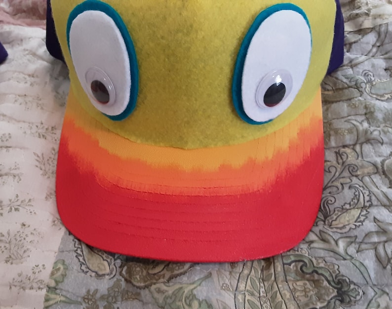 May include: A yellow felt hat with orange and red trim. The hat has two large white eyes with black pupils.