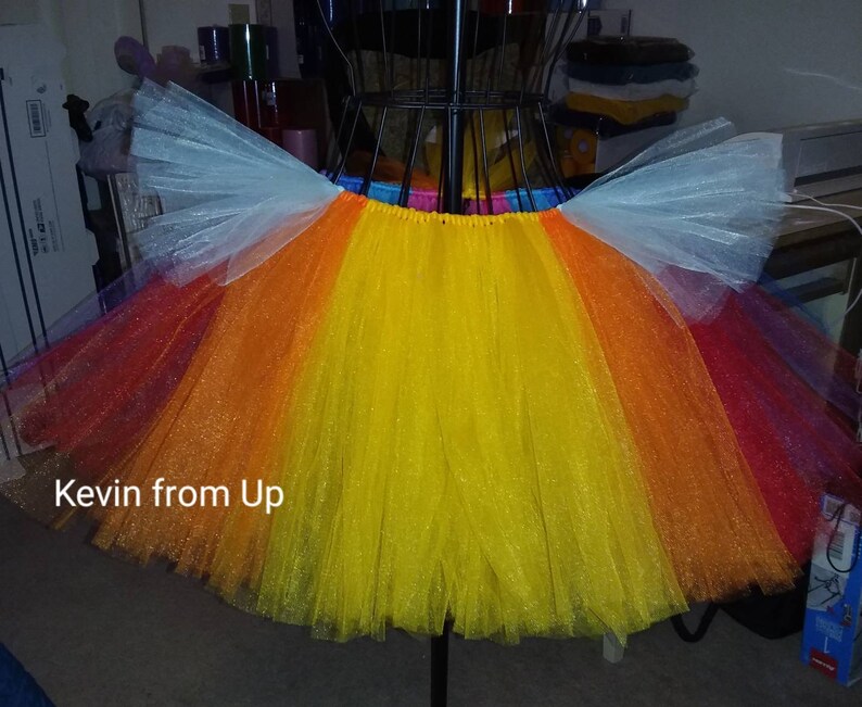 May include: A rainbow tulle tutu with white tulle wings. The tutu has a yellow, orange, red, and purple layers. The text "Kevin from Up" is written on the tutu.