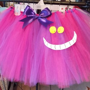 May include: A pink and purple tulle tutu with a purple bow and a white Cheshire Cat face with yellow eyes.