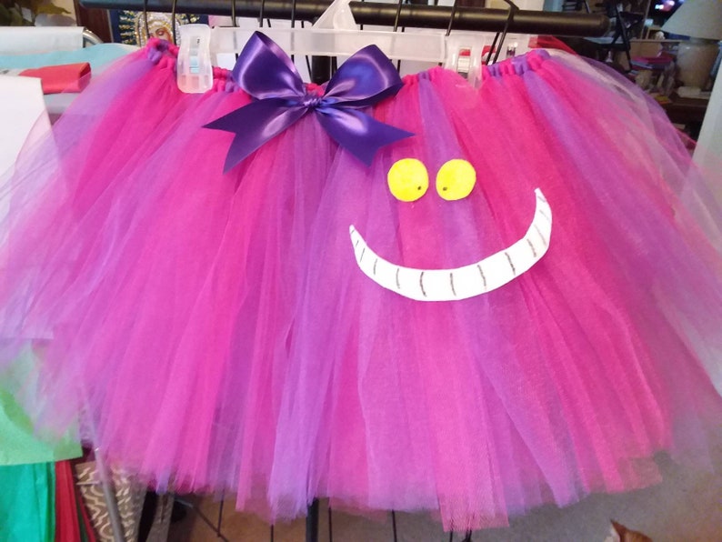 May include: A pink and purple tulle tutu with a purple bow and a white Cheshire Cat face with yellow eyes.
