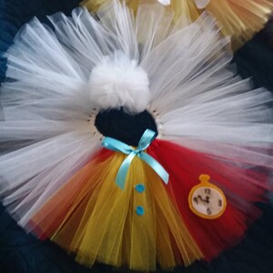 May include: A white tulle tutu with a red, yellow, and orange layered skirt. The tutu has a blue bow and two blue buttons. A felt clock with Roman numerals is attached to the red portion of the skirt.