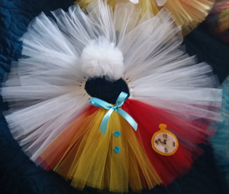 May include: A white tulle tutu with a red, yellow, and orange layered skirt. The tutu has a blue bow and two blue buttons. A felt clock with Roman numerals is attached to the red portion of the skirt.