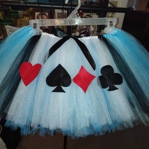 May include: A blue and black tulle tutu skirt with a white glitter overlay. The skirt has four felt appliques of playing card suits: a red heart, a black spade, a red diamond, and a black club.