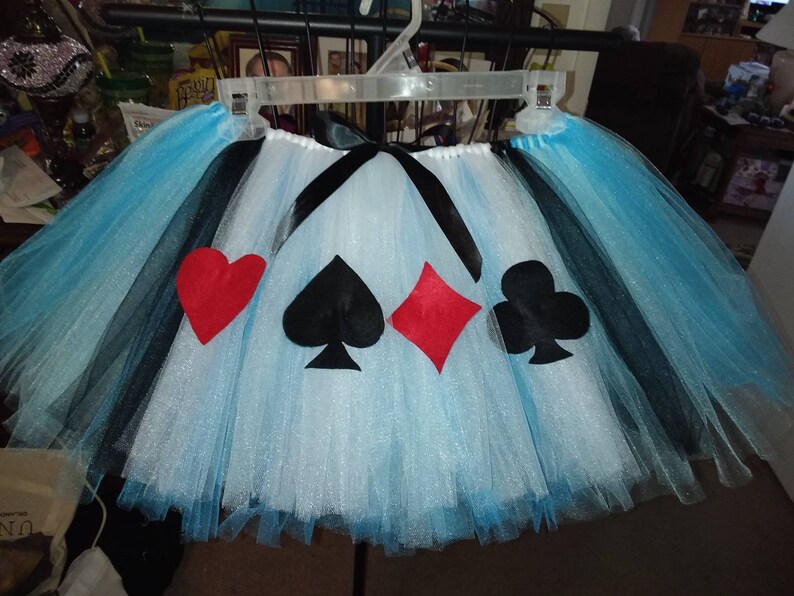 May include: A blue and black tulle tutu skirt with a white glitter overlay. The skirt has four felt appliques of playing card suits: a red heart, a black spade, a red diamond, and a black club.