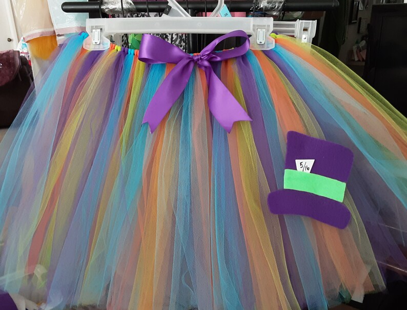 May include: A rainbow tulle tutu with a purple ribbon and a purple felt hat with a green band and the text "5/16" on it.