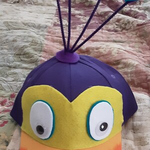 May include: A purple felt hat with a yellow beak and large white eyes. The hat has a rainbow-colored brim and a purple felt peacock tail with blue feathers.