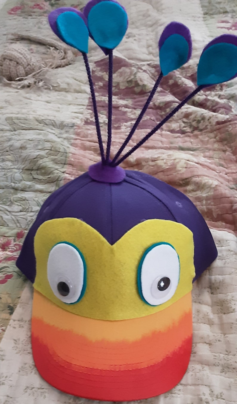 May include: A purple felt hat with a yellow beak and large white eyes. The hat has a rainbow-colored brim and a purple felt peacock tail with blue feathers.