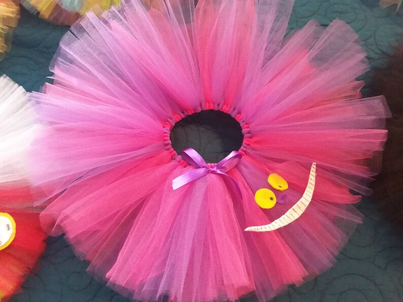 May include: A pink and purple tulle tutu with a white felt Cheshire Cat grin and yellow felt eyes.