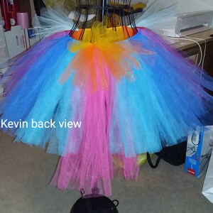 May include: A multi-layered tulle skirt with a rainbow color scheme. The skirt is made of layers of pink, blue, and orange tulle, with a yellow waistband. The skirt is hanging on a hanger and the text "Kevin back view" is visible at the bottom of the image.