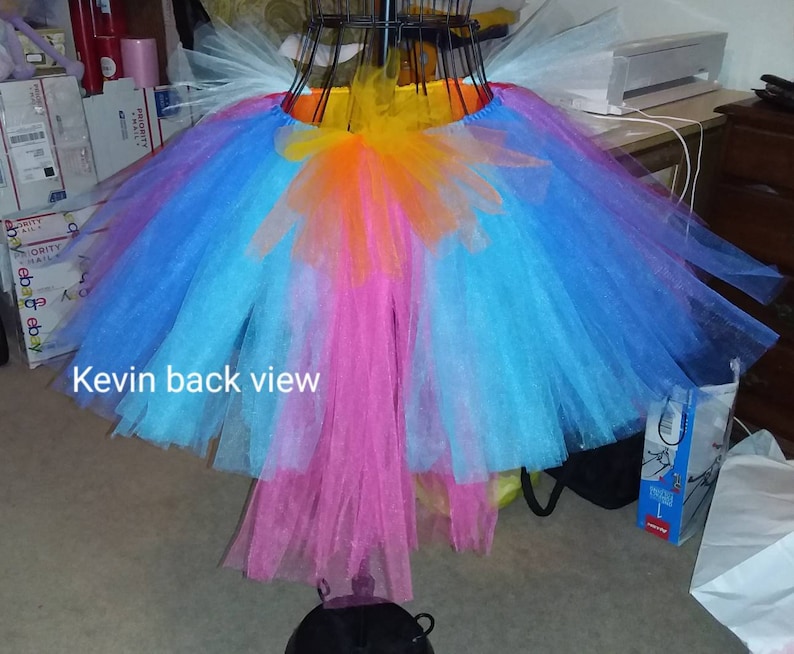 May include: A multi-layered tulle skirt with a rainbow color scheme. The skirt is made of layers of pink, blue, and orange tulle, with a yellow waistband. The skirt is hanging on a hanger and the text "Kevin back view" is visible at the bottom of the image.