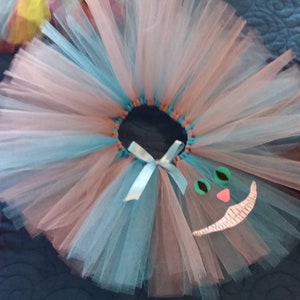 May include: A light blue and pink tulle tutu with a white Cheshire Cat face appliqu&eacute;. The face has green eyes, a pink nose, and a white smile.