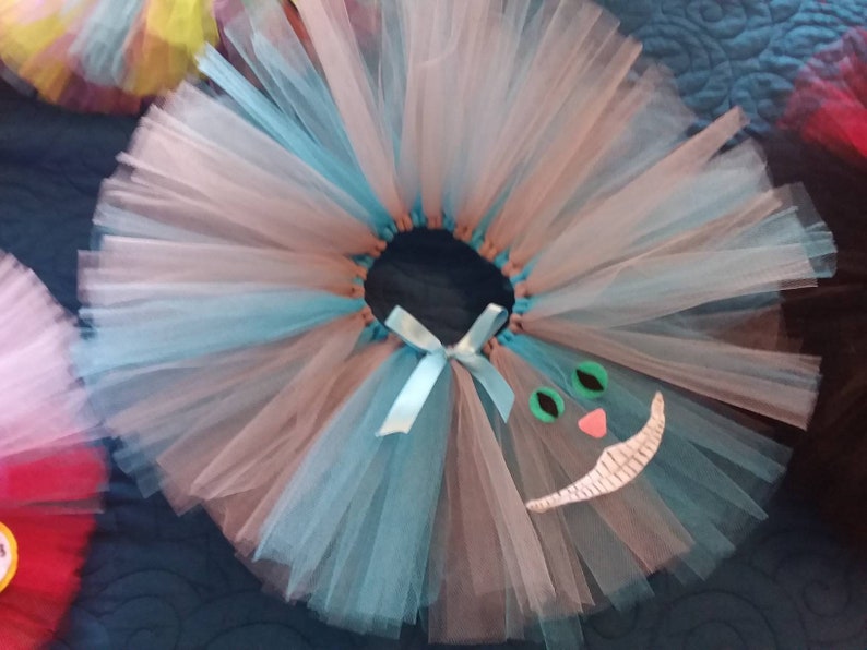 May include: A light blue and pink tulle tutu with a white Cheshire Cat face appliqu&eacute;. The face has green eyes, a pink nose, and a white smile.