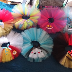 May include: Six colorful tulle tutus with different Alice in Wonderland character appliques. The tutus are arranged in a circle on a blue surface. The characters include the Queen of Hearts, the Mad Hatter, the Cheshire Cat, and the White Rabbit.
