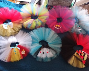 May include: Six colorful tulle tutus with different Alice in Wonderland character appliques. The tutus are arranged in a circle on a blue surface. The characters include the Queen of Hearts, the Mad Hatter, the Cheshire Cat, and the White Rabbit.