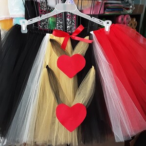 May include: A multi-colored tulle skirt with black, white, gold, and red layers. The skirt has two red felt hearts attached to the front. The skirt is hanging on a white hanger.
