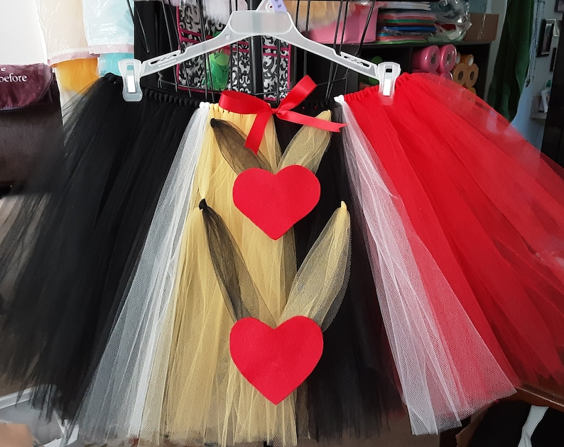 May include: A multi-colored tulle skirt with black, white, gold, and red layers. The skirt has two red felt hearts attached to the front. The skirt is hanging on a white hanger.