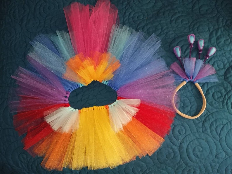 May include: A rainbow tulle tutu skirt with a matching headband featuring purple and blue tulle flowers. The skirt is made of multiple layers of tulle in various colors, including red, orange, yellow, green, blue, and purple.