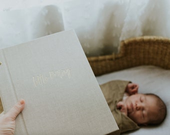May include: A light gray linen baby memory book with the text "Little Moments" embossed in gold. The book is being held by a hand. A baby is sleeping in a wicker basket.