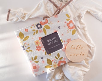 May include: A cream-colored baby gown with brown trim, a floral-patterned keepsake book, and a wooden disc with the words "hello world" engraved. The book has a square opening and the words "MODERN KEEPSAKE" printed on it.
