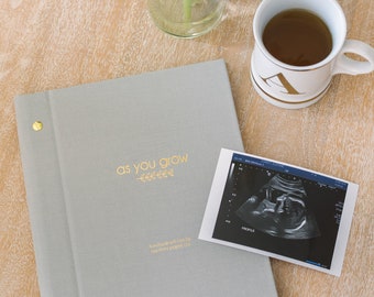 May include: A gray fabric-covered journal with gold lettering that reads "as you grow" and a small floral design. The journal is on a wooden table with a cup of tea and a sonogram image.
