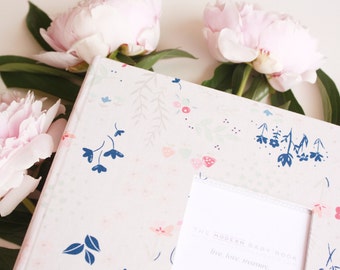 May include: A white baby book with a floral design and a frame that says "The Modern Baby Book Live Love Treasure". The book is surrounded by pink peonies.