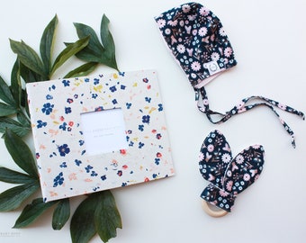 May include: A floral baby album with a window for a photo, a floral baby hat with ties, and a floral teething ring with a wooden ring.