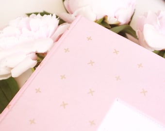 May include: A pink notebook with a gold star pattern. The notebook is lying on a white surface with pink flowers.