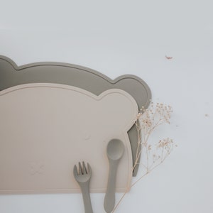 May include: Two silicone placemats in the shape of a bear, one beige and one gray, with a beige spoon and fork on top.