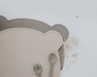 May include: Two silicone placemats in the shape of a bear, one beige and one gray, with a beige spoon and fork on top.
