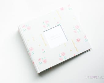 May include: A white photo album with a floral pattern and a rectangular window for a photo. The album has a delicate floral design with pink and green flowers.