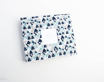 May include: A square photo album with a geometric pattern of blue, gray, and teal triangles on a white background. The album has a square cutout in the center, with text visible inside. The overall theme is a baby or family keepsake.