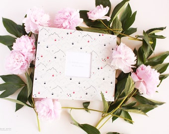 May include: A white baby book with a mountain and cable car pattern. The book is surrounded by pink peonies and green leaves. The text on the book reads "The Modern Baby Book".
