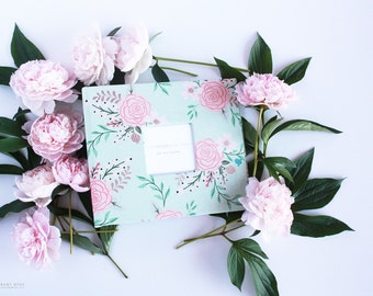 May include: A light green scrapbook with a floral design featuring pink and white flowers. The scrapbook has a white window in the center with the text "Our Modern Baby Book" and "The Love Story Begins". The scrapbook is surrounded by pink peonies and green leaves.