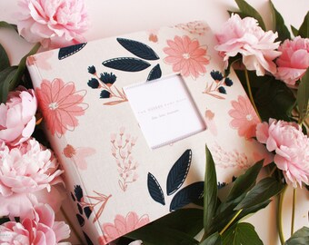 May include: A white fabric photo album with a floral design featuring pink and blue flowers. The album has a clear window on the front with the text "The Wedding Album" and "Our Love Story".