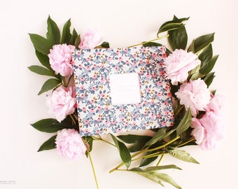 May include: A floral patterned notebook with a white card inside. The notebook is surrounded by pink peonies and green leaves.