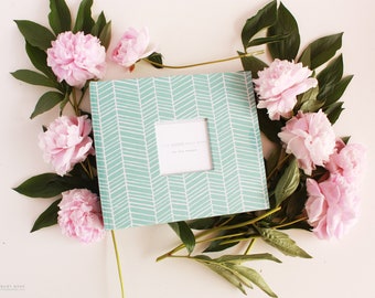 May include: A teal blue baby book with a white chevron pattern and a white window with the text "The Modern Baby Book" and "Your Baby's Journey". The book is surrounded by pink peonies and green leaves.