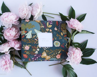 May include: A teal blue baby book with a floral pattern and a white frame with the text "The Modern Baby Book" and "Our Baby Journey". The book is surrounded by pink peonies and green leaves.