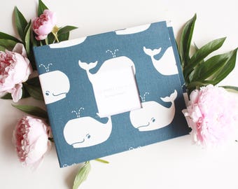 May include: A blue fabric photo album with a white whale pattern. The album has a white window for a photo. The album is surrounded by pink peonies.