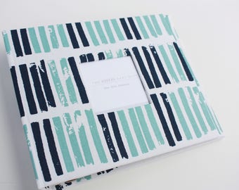 May include: A white fabric photo album with a blue and teal striped pattern. The album has a window for a photo with the text "The modern family album for all your memories".