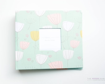 May include: A light green baby photo album with a white frame and a floral design featuring pink and yellow flowers. The text "Our Story Starts Here" is printed inside the frame.