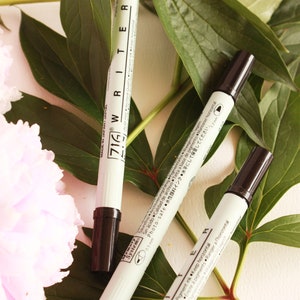 May include: Three white markers with black caps and black writing. The markers have Japanese text on them that reads "716 Writer" and "Photo-safe Ink".