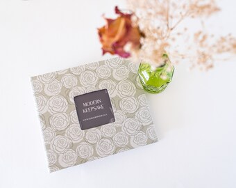 May include: A beige photo album with a white floral pattern and a window showing the text "MODERN KEEPSAKE".