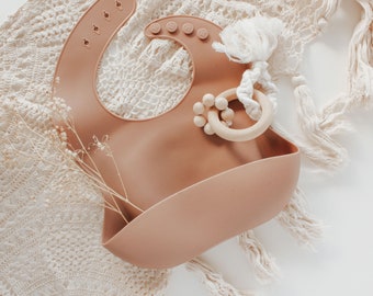 May include: A light brown silicone bib with a pocket for catching food. The bib is on a white crocheted surface with a wooden teether with beads.