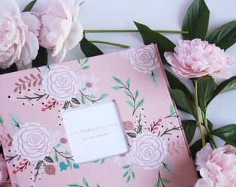 May include: A pink baby memory book with a floral design and a white window for a photo. The text "The Modern Baby Book" and "The love story" is printed on the cover.