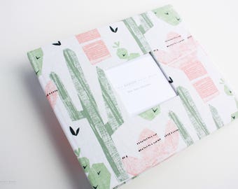 May include: A white fabric photo album with a cactus and flower design. The album has a window for a photo and the text "The moments we share, the love we treasure".