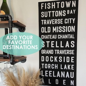 May include: A rectangular black canvas sign with white text listing locations including Fishtown, Suttons Bay, Traverse City, Old Mission, Chateau Chantal, Stellas, Grand Traverse, Dockside, Torch Lake, Leelanau, and Alden, measuring 16 inches by 36 inches.